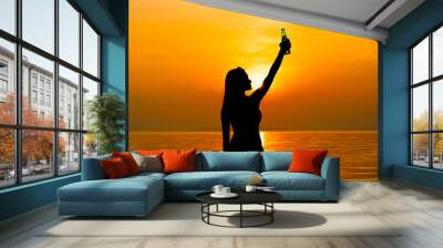 Silhouette of woman raising her hand with beer bottle at the beach in sunset Wall mural