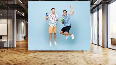 Smilng happy male friends playing with water guns and jumping in blue isolated background for Songkran festival in Thailand and southeast Asia Wall mural