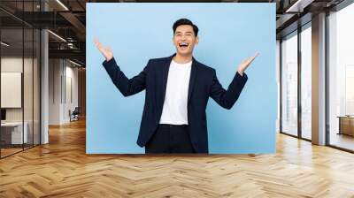 Surprised worry free handsome Asian man in semi formal clothes smiling and opening hands in light blue isolated studio background Wall mural