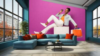 Trendy fashionable African American woman holding colorful shopping bags and credit card sitting on stool in pink color studio isolated background Wall mural