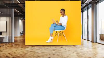 Young African American woman sitting and using tablet computer on isolated yellow background Wall mural