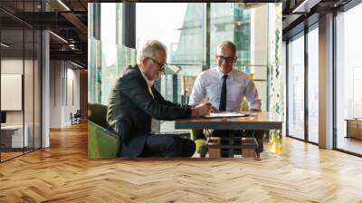 Picture of two business man discussing business on a table.  The young man is wearing a white shire with black tie and glass. The older man wear a black suit and sunglass. They are in a hotel lobby. Wall mural