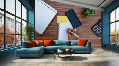 Writing on a notepad while working from home. A lap top, a mobile, calculator and plant are also on display on this brown striped working table. Wall mural