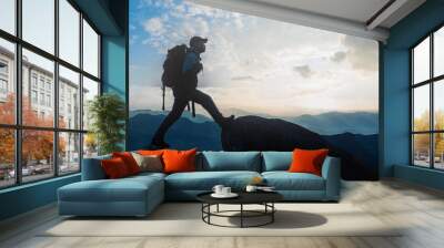 Low angle view of Asian male hikers climbing mountain at sunset rays over the clouds with trekking poles on cliff edge on top of rock mountain, Successful summit concept image. Wall mural