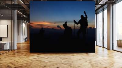 Silhouette of Asian teamwork standing raised hands with trekking poles and Backpack and other camping gear on cliff edge on top of rock mountain with at sunset rays over the clouds background,  Wall mural