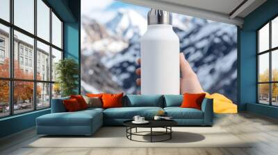 A person holds a white insulated water bottle against a backdrop of snowy mountains and blue skies. Wall mural