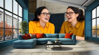 two happy female colleagues laughing together at work, positive teamwork concept, modern office setting, yellow shirts, smiling women, creative collaboration, business partners. Wall mural