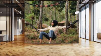 Pensive smiling young woman with headscarf resting in comfortable hammock at green garden. Wall mural