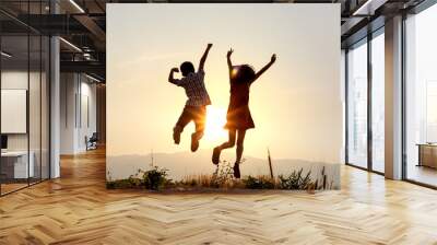 Silhouette of happy little boy and girl jumping playing on mountain at sunset time Wall mural