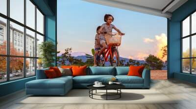 two children playing riding bike in park at sunset sky background Wall mural