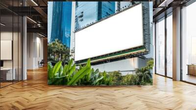 A large blank billboard situated in front of skyscrapers surrounded by foliage, ready for advertising, against a clear sky in an urban setting. Wall mural