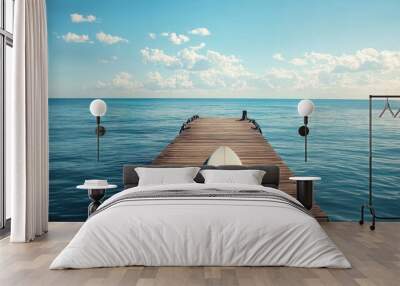 A serene wooden pier extends into a calm ocean under a clear sky, with a surfboard resting on it. Wall mural