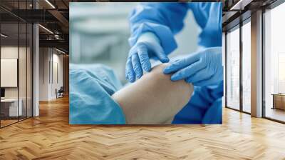 A healthcare professional in blue gloves bandages a patient's knee in a clinical setting, emphasizing care and medical attention. Wall mural