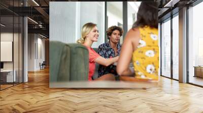 Candid shot of three multi-ethnic friends sitting together at table in bright trendy cafe serving fair-trade coffee wearing fashionable clothing Wall mural