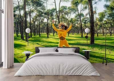 A girl in a yellow tracksuit jumping for joy in the park. Wall mural