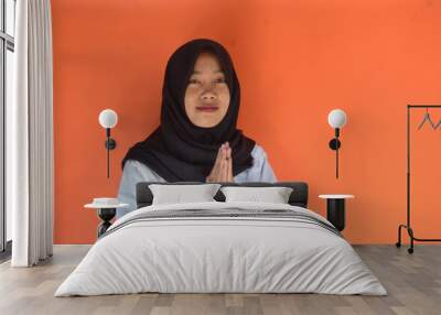 Portrait of smiling beautiful young Asian Muslim woman wearing hijab and white shirt standing with Eid greeting gesture and welcoming Ramadan isolated on orange studio background Wall mural