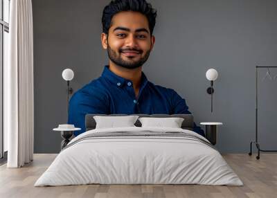 A handsome Indian man in a blue shirt stands with his arms crossed against a grey background. Wall mural