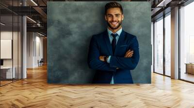 A happy young businessman in a blue suit with arms crossed, standing against a gray wall. Wall mural