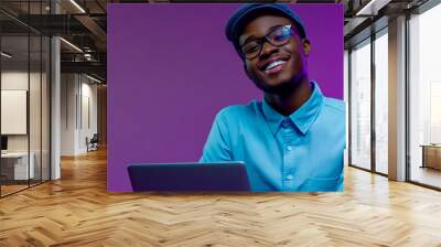 A happy young man with a laptop on a purple background, wearing a blue shirt and cap. Wall mural