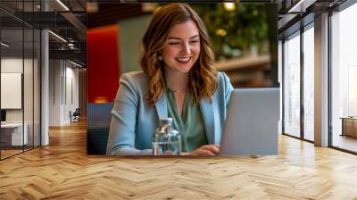 A young woman in professional attire is sitting at her laptop, smiling as she pours water from the glass on her desk. Wall mural