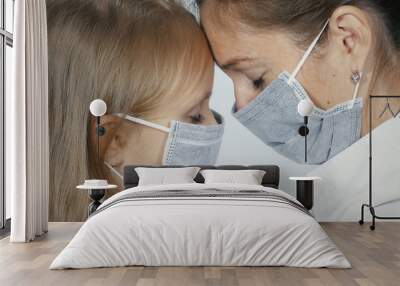 Close-up of female doctor wearing medical mask sitting face to face with little blonde girl in medical mask. The doctor calms the baby and hugs. Wall mural