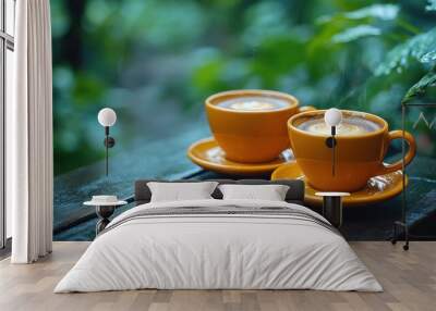 Two steaming cups of coffee with latte art on a wooden table with coffee beans in front of a green leafy background. Wall mural