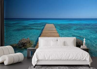 Wooden pier extending into a turquoise blue ocean with clear water and two large rocks. Wall mural