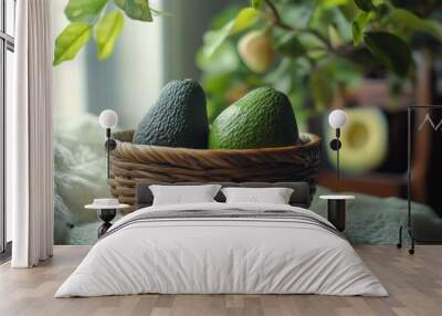 Two ripe avocados in a wicker basket on a white cloth with a green plant and a blurred background. Wall mural