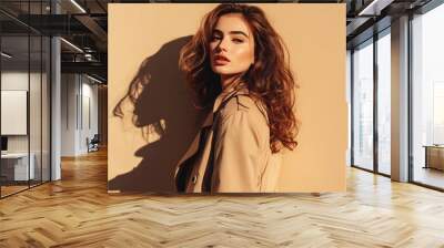 Beautiful young woman with long brown hair in a beige coat posing against a beige background. Wall mural