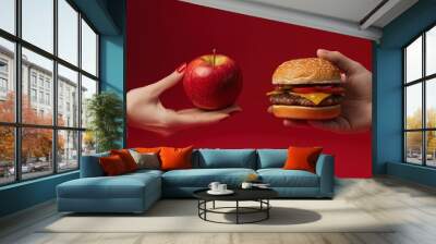 Female hand holding apple and burger on red background, showing healthy food versus fatty hamburger or fast food bun. Concept with space for text in the center. Wall mural