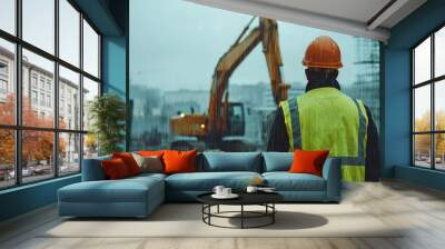 The back of an engineer wearing a yellow vest and orange helmet stands in front of an excavator on a construction site, with heavy rain falling from the sky. It is a rainy day with a blurred backgroun Wall mural