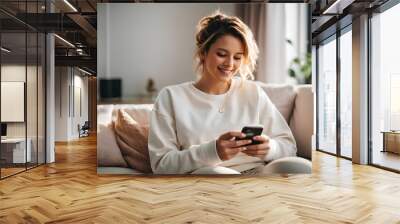 A smiling girl is sitting on the sofa with a phone in her hands. Wall mural