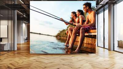 Group of friends sitting on pier by the lake and fishing.They joying in beautiful summer sunset. Wall mural