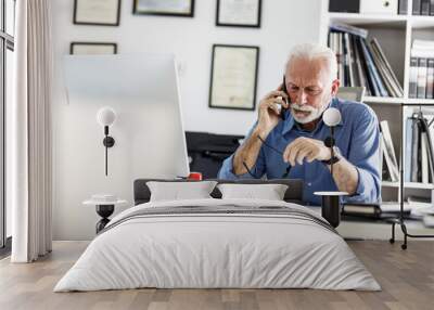 Senior business manager sitting at the desk in his office and speaking on smart phone.Looking serious and trying to solve problem. Wall mural