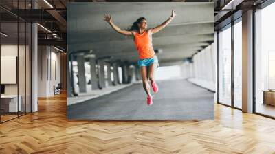 Young female runner jumps on the street,expressing positive emotion.She made her goal of the day.Fitness and workout concept. Wall mural