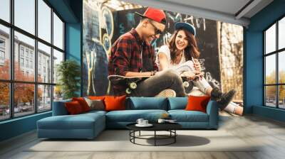 Young teen couple sitting by the wall and hangout at the street .Embrace each other and laughing . Wall mural