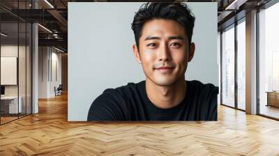 An attractive Asian man with a charming smile, posing with his arms folded against a crisp white background. He is casually dressed in a black t-shirt, exuding a relaxed and approachable vibe Wall mural