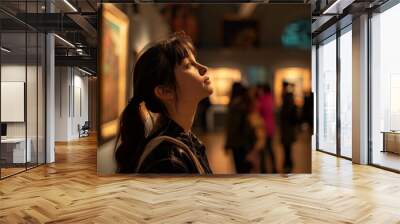 A woman is looking at a painting in a museum. She is wearing a black jacket and has her hair in a ponytail. There are other people in the room, some of whom are looking at the same painting Wall mural