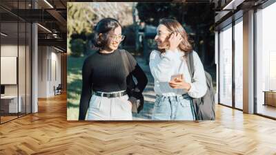 Two young woman students small talking while going to class at the college during a sunny day holding backpacks and having fun. Back at school concept. Multi cultural students at the university Wall mural