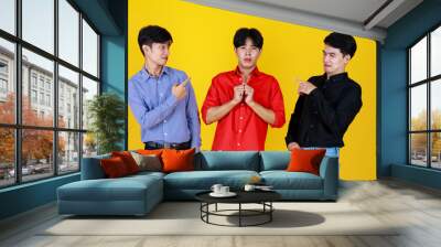 3 adults asian men close friends team, 2 men finger pointing to 1 of team who feel confused look like don't understand with something. Shooting in studio with isolated yellow background. Wall mural