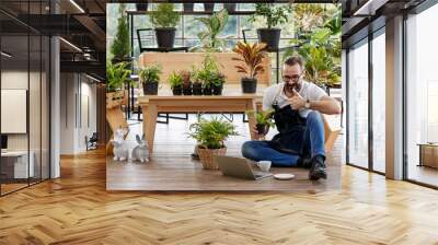 A beautiful bearded man sitting and using laptop notebook computer to sell plant in home indoor garden. Online marketing and live stream seller concept Wall mural