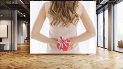 A long brown hair female wearing pink sleeveless dress stand hiding red gift box with white ribbon behind her back Wall mural