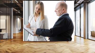 Adults caucasian businessman boss and staff woman seeing a chart graph on report paper together as present by women staff to boss near window. Woman look easy dress with white shirt and blue jean Wall mural