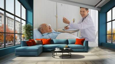 An old fat asian man with white hair wearing eyeglasses and light blue shirt lay down on the bed in hospital room while male doctor with short black hair wearing white lab coat smiling checking  Wall mural