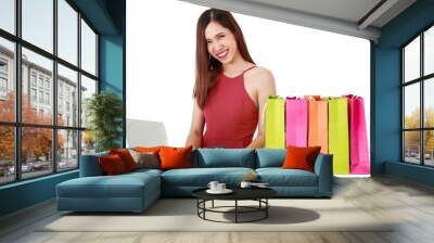 Asian beautiful smiley woman wearing fashion cloth, using laptop for doing online shopping payment with happiness. There are colorful paper bags on table, sitting on isolated white background cutout. Wall mural