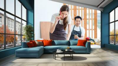 Asian professional female carpenter in apron holding pencil using square ruler measuring wood stick on wooden table with equipment in home decoration building workshop while male colleague take break Wall mural