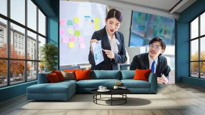 Asian professional successful female businesswoman lecturer presenter in formal suit standing smiling at graph chart data answer questions to male colleague in company office meeting room. Wall mural