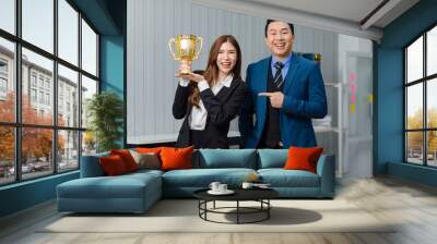 Asian professional successful male businessman ceo entrepreneur mentor  in formal business suit standing smiling pointing young female businesswoman holding golden cup trophy award for best employee Wall mural