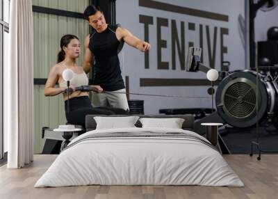 Asian strong young fit male trainer helping teaching female muscular fitness athlete model in sexy sport bra and legging sitting using cable pulling machine working out training exercising in gym Wall mural