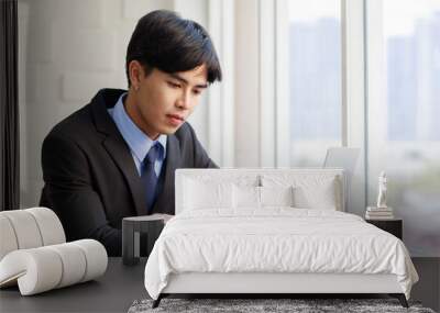 Asian young businessman are smart and handsome wearing black suit sitting while typing on keyboard at laptop near window in office at the morning Wall mural